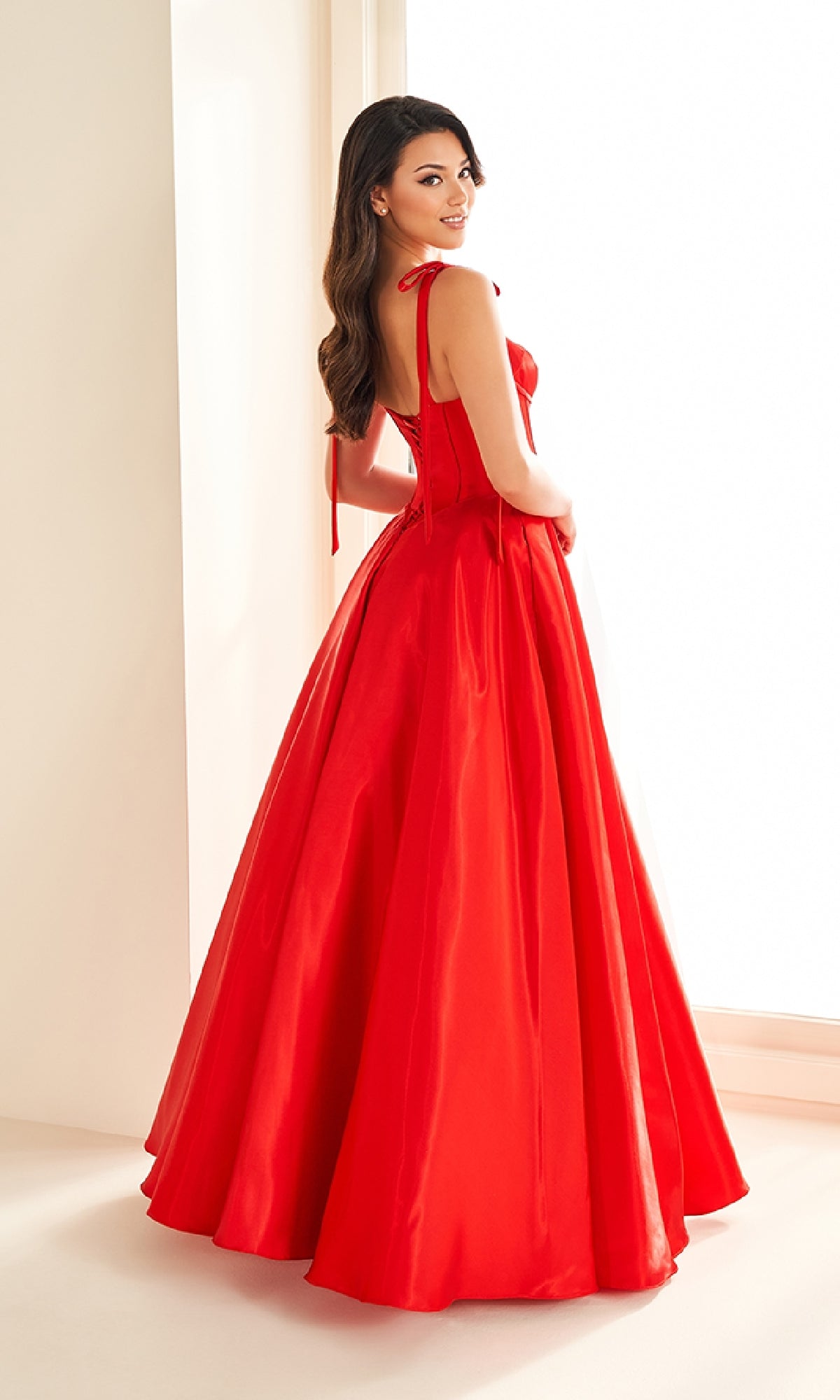 A woman in the Ellie Wilde EW36210 Shoulder-Bows Prom Ball Gown stands by a window, glancing over her shoulder with a smile.